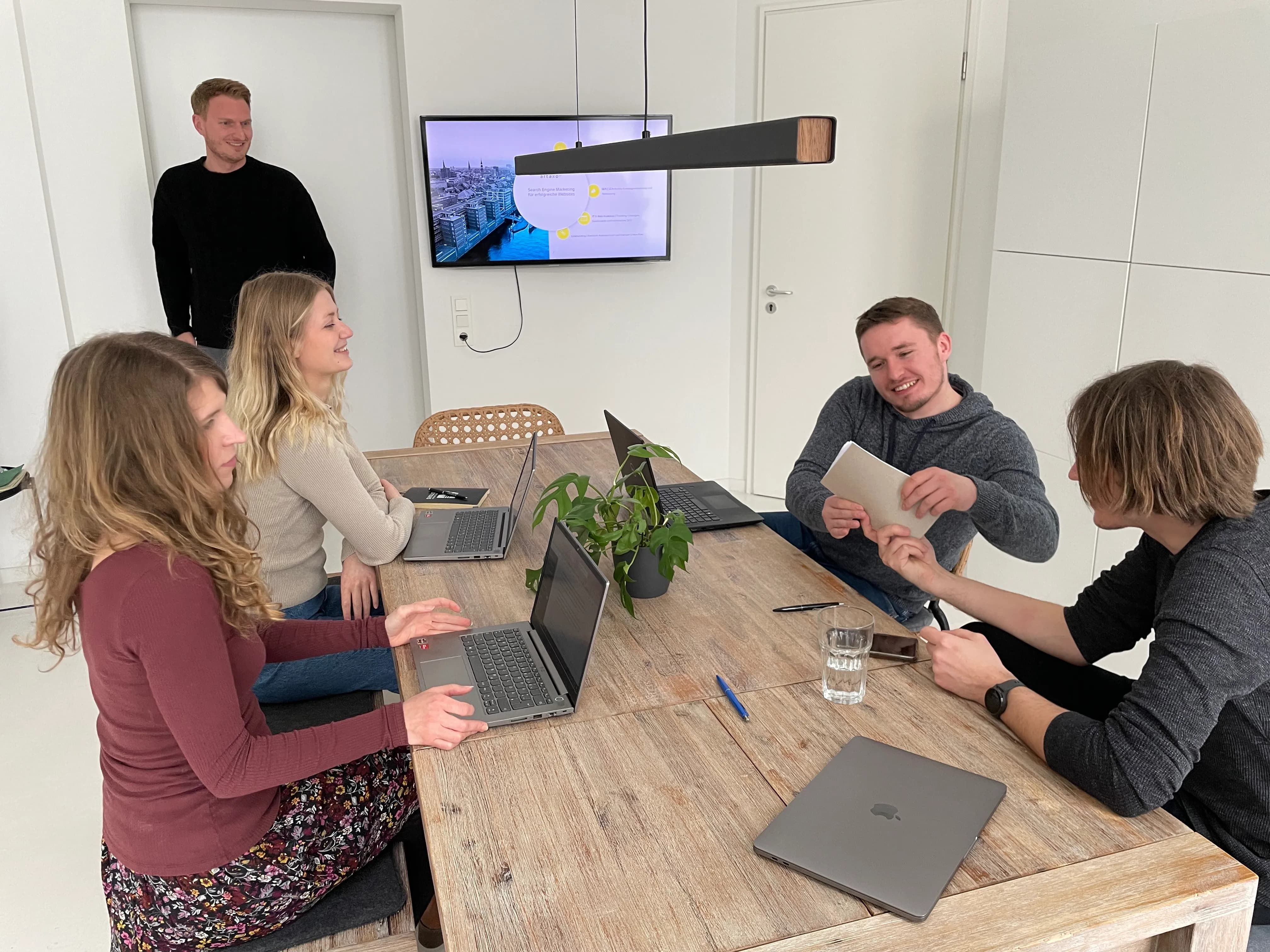 Ein Team-Meeting im Konferenzraum des Büros. Die Frauen und Männer unterhalten sich über die Präsentation, die hinten auf dem Fernseher präsentiert wird.