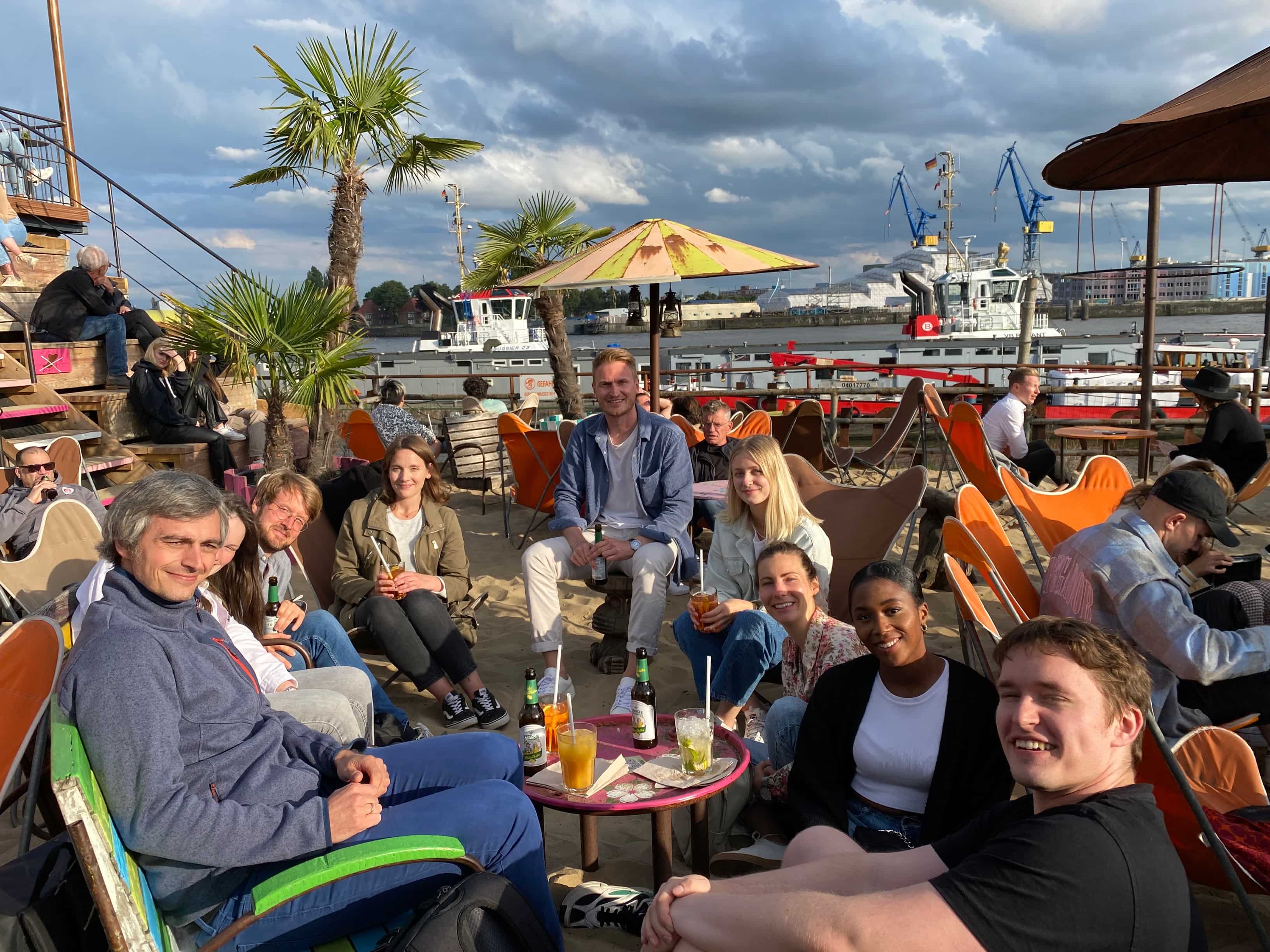 After-work get-together at a beach bar on the Elbe. Colleagues sit in a circle in the sand and on chairs, looking into the camera with summer drinks on the table.